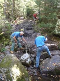 Crew using a winch, cable, and bars to guide a rock step into place on the Mount Kinsman Trail