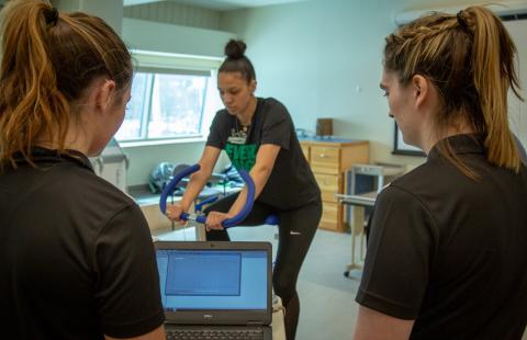 Two female students looking at a laptop screen face a female student on a Stairmaster.