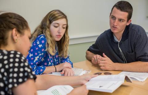 Three Plymouth State University students sit at a round table looking at written materials and talking amongst themselves.