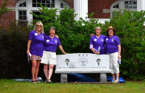 Four women wearing matching purple shirts stand in front of a granite bench